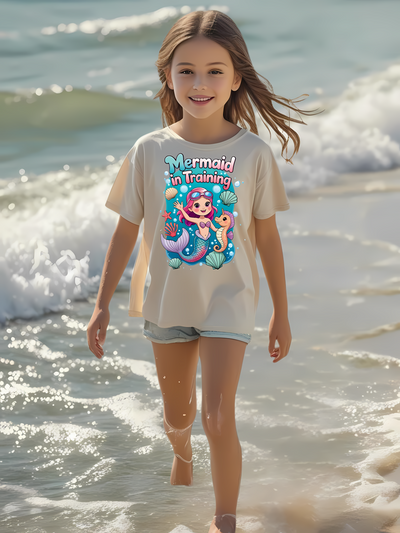 Young girl wearing a 'Mermaid in Training' graphic t-shirt at the beach