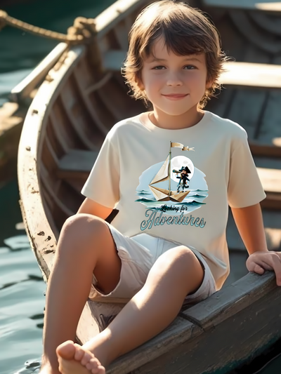 Child sitting in a wooden boat wearing a crew neck t-shirt with a pirate on a paper folded sailboat design and 'Looking for Adventure' text.